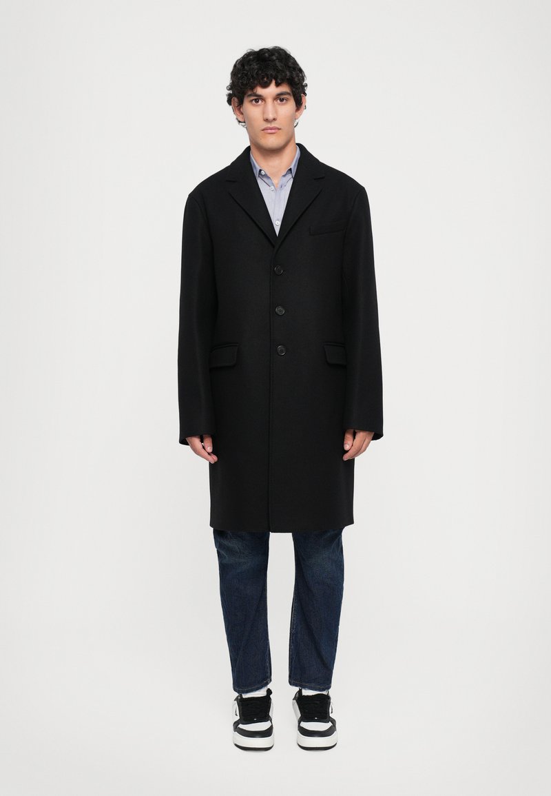 Black wool coat with a notch lapel, three button closure, and two front pockets, worn over a light blue shirt and dark jeans.