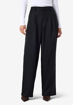 Femme portant un pantalon noir taille haute à jambes larges avec passants de ceinture, associé à un haut blanc à manches longues et des chaussures marron pointues.