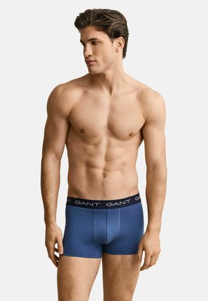 Muskulöser Mann mit kurzen dunklen Haaren, der blaue GANT-Boxershorts trägt und vor einem einfarbigen hellen Hintergrund steht, dabei nach links blickt.