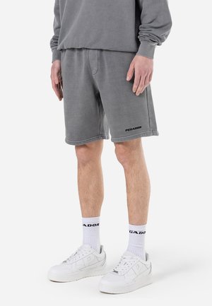 Graue Baumwollshorts mit Seitentaschen, überstehenden Nähten und einem schwarzen Logo am linken Bein. Kombiniert mit weißen Sneakers und Socken mit Logo-Akzenten.