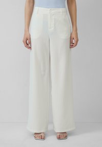 Pantalon blanc à jambes larges avec des rayures fines bleu clair, présentant une taille plate, des passants de ceinture et des poches latérales. Porté avec des sandales à talons argentées.
