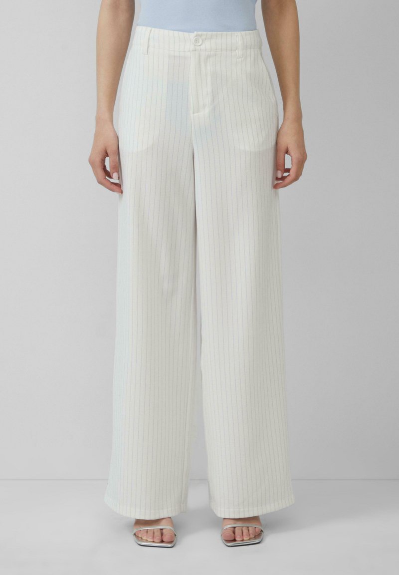 Pantalon blanc à jambes larges avec des rayures fines bleu clair, présentant une taille plate, des passants de ceinture et des poches latérales. Porté avec des sandales à talons argentées.