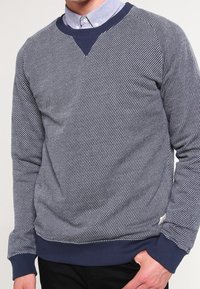 Navy blue crewneck sweater with diagonal white stripes, ribbed cuffs, and a textured finish. Layered over a light blue collared shirt.