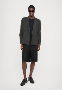 Γκρίζα διπλή σακάκι πάνω από ένα σκούρο πουκάμισο, συνδυασμένο με μαύρες βερμούδες και loafers. Τα αξεσουάρ περιλαμβάνουν μαύρα γυαλιά ηλίου.