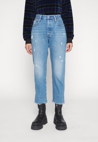 Vaqueros desgastados de color azul claro con corte recto, que presentan detalles de dobladillo desflecado y varios pequeños rasguños. Combinados con botas negras de suela gruesa.