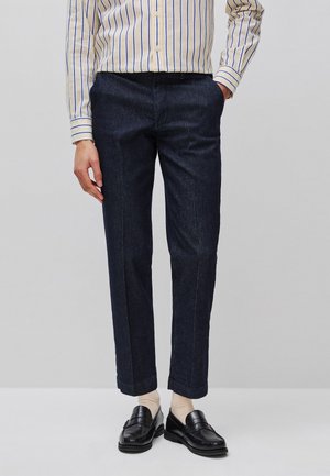 Mężczyzna ubrany w ciemnoniebieskie spodnie o prostych nogawkach, beżowe skarpetki, czarne mokasyny i kremową koszulę z niebieskimi pionowymi paskami, stojący na jednolitym tle.