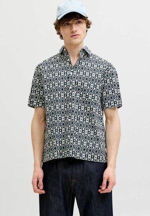 Mladić koji nosi kratku crno-bijelu uzorkovanu košulju, tamne traperice i svijetloplavu kapu na jednobojnoj pozadini.