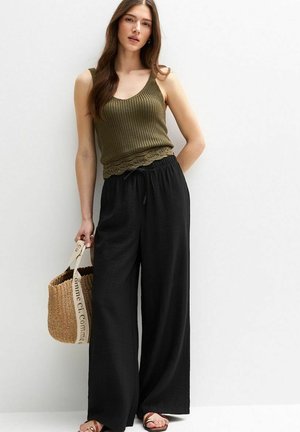 Olivenfarbener, gerippter Tanktop mit Spitzenbesatz, kombiniert mit schwarzen, weit geschnittenen Hosen. Das Model hält eine geflochtene Strohtasche mit bedruckten Trägern.