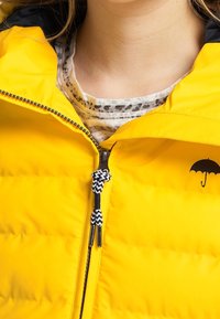 Gul puffjacka med dragkedja i svart och mönstrad dragring. Har en liten svart paraplylogotyp på vänster sida.