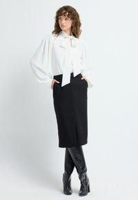 Chemisier blanc avec un col noué, des manches bouffantes, et une jupe crayon noire jusqu'au genou. Bottes en cuir texturé noires, montantes jusqu'aux genoux.