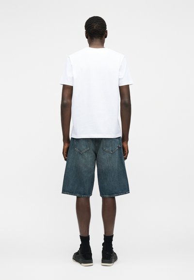 Άνδρας που στέκεται με την πλάτη στην κάμερα, φοράει απλό λευκό T-shirt, μακριά τζιν σορτς, μαύρες κάλτσες και μαύρα αθλητικά παπούτσια σε λευκό φόντο.