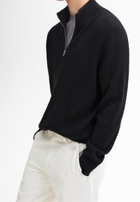 Black ribbed knit sweater with a quarter-zip design, textured fabric, and a high collar, paired with light-colored trousers.