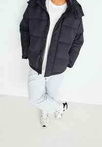 Veste matelassée noire à fermeture éclair avec capuche, présentant un design matelassé et des poignets élastiques, portée sur une chemise blanche et un jean bleu clair.