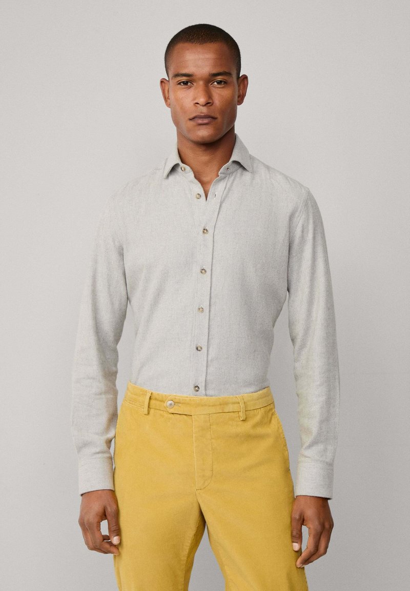 Camisa de manga larga de color gris claro con cuello abotonado y botones marrones, combinada con pantalones amarillos mostaza, que presentan una textura suave.