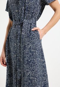 Marineblaues Kleid mit floralem Muster, kurzen Ärmeln, Knöpfen vorne, tailliertem Gürtel und Seitentaschen. Glatte Stoffstruktur.