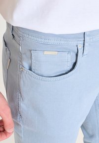 Pantalones de mezclilla azul claro que cuentan con un bolsillo frontal con un detalle plateado y una pequeña etiqueta de marca. La tela tiene una textura suave.