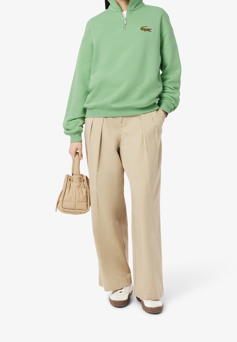 Personne portant un sweat-shirt zippé vert clair avec un logo crocodile, un pantalon large beige, des baskets blanches, et tenant un petit sac à main beige.