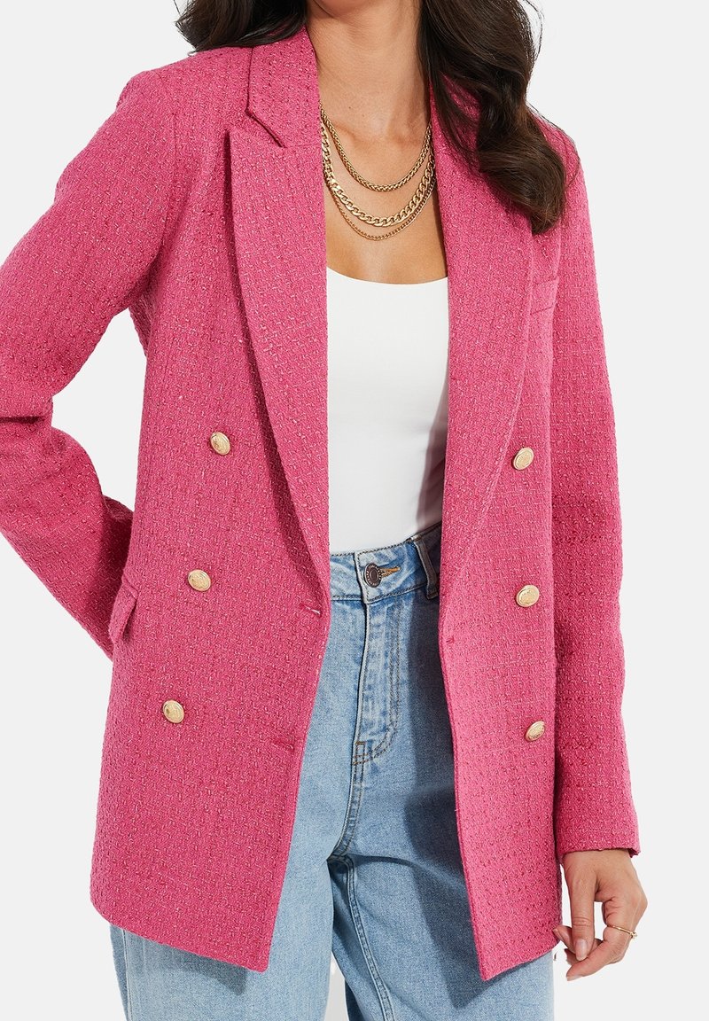 Blazer double boutonnage rose fuchsia en tissu texturé, avec boutons en métal doré, revers crantés et poches avant, assorti à un haut blanc.