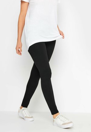 Mustat legginssit sileällä pinnalla, nilkkoihin ulottuvalla muotoilulla, yhdistettynä valkoiseen ylisuurikokoiseen t-paitaan ja valkoisiin tenniskenkiin.