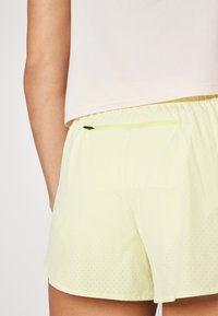 Pantalones cortos amarillos claros con textura perforada, cinturilla elástica y un bolsillo trasero con cremallera de contraste en verde.