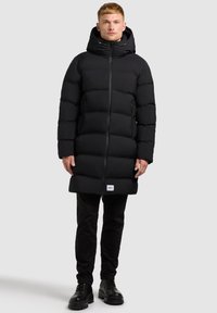 Polstret sort parka med hætte, der har en fuld lynlås, sidelommer og et mærke ved kanten. Bæres over sorte bukser og støvler.