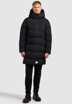 Padded black parka with a hood, featuring a full zipper, side pockets, and a label at the hem. Worn over black pants and boots.