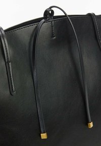 Gros plan sur un sac tote en cuir noir montrant de fines poignées de shoulder avec des embouts métalliques dorés en forme de carré, sur un fond uni.