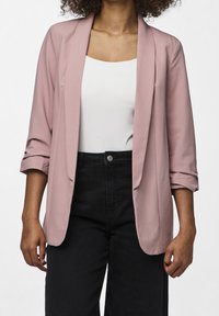 Blazer rose pâle à texture douce, avec une coupe ouverte sur le devant et des manches retroussées, assorti d'un haut blanc et d'un pantalon noir taille haute.