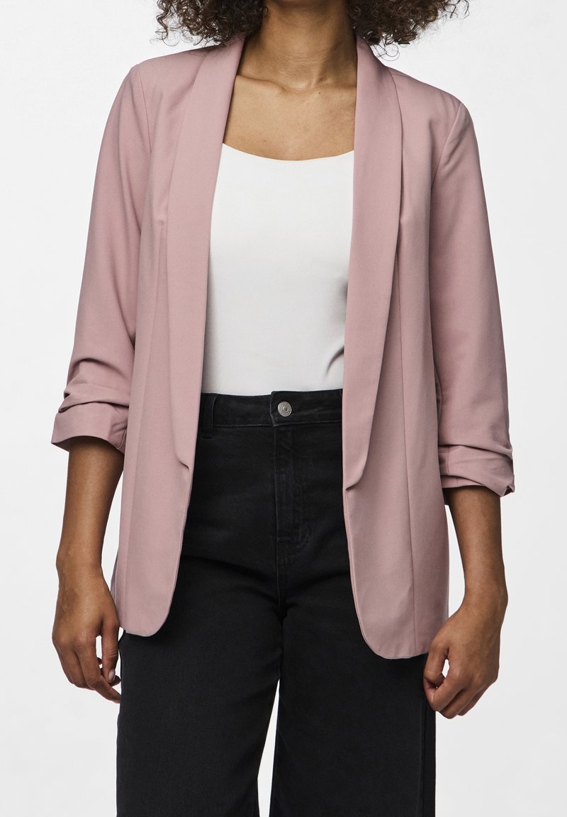 Blazer rose pâle à texture douce, avec une coupe ouverte sur le devant et des manches retroussées, assorti d'un haut blanc et d'un pantalon noir taille haute.