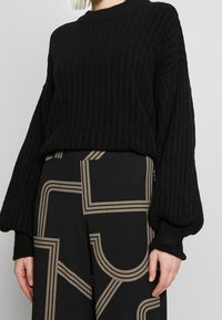 Svart ribbad tröja med vida långa ärmar, i kombination med mönstrade svarta och gyllene byxor som har geometriska linjer och former.