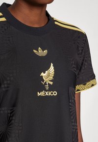 Svart idretts skjorte med gull Adidas-logo, emblem av en fugl, og teksten "MÉXICO"; strukturert design med mønstrede ermer og trim.