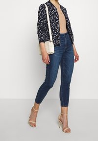 Kvinna som bär en marinblå blommig jacka, beige ribbad topp, mörka skinny jeans, beige högklackade sandaler och bär en vit flätad axelremsväska.