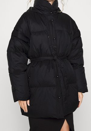 Personne portant une doudoune noire oversize avec un col haut, des boutons-pression et une ceinture nouée à la taille à l'avant.