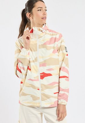 Femme ajustant le col d'une veste à capuche zippée à l'avant, camouflage beige, rose et rouge, debout devant un fond clair uni.