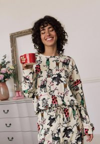 Conjunto de ropa de casa de algodón que presenta una tela color crema con patrones coloridos de renos y perros, mangas largas y cintura elástica. Sosteniendo una taza roja.