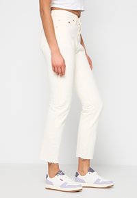 Jeans de denim blanco con cintura alta, pierna recta y dobladillo deshilachado. Llevados con zapatillas blancas y lavanda.