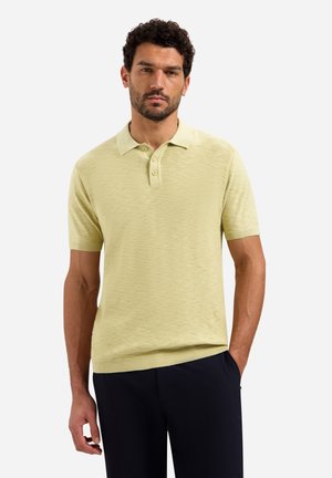 Man met krullend haar, gekleed in een lichtgeel poloshirt met korte mouwen en een donkere broek, staand met één hand in zijn zak tegen een witte achtergrond.