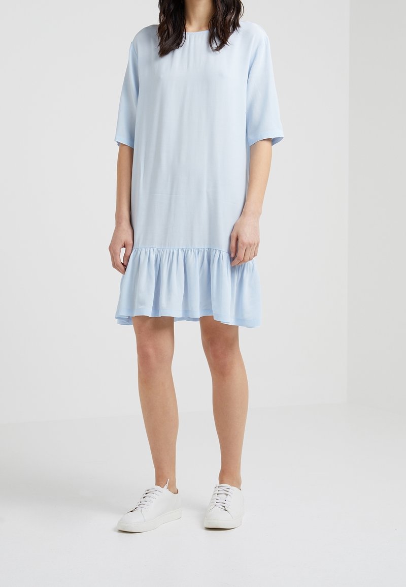 Femme portant une robe bleu clair longueur genou avec un ourlet à volants et des manches aux coudes, associée à des baskets blanches à lacets en dentelle.