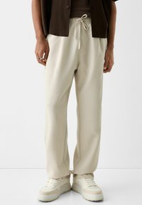 Béžové tepláky se stahovací šňůrkou v pase, s rovnými nohavicemi a jemným postranním švem. Kombinovány s světlými teniskami.