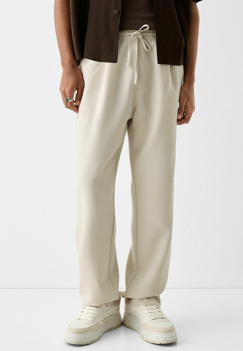 Béžové tepláky se stahovací šňůrkou v pase, s rovnými nohavicemi a jemným postranním švem. Kombinovány s světlými teniskami.