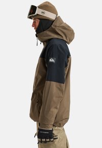 Braune und marineblaue wasserdichte Jacke mit Kapuze, Seitentaschen, Logopatch am Arm, getragen mit schwarzen Handschuhen. Strukturierte Strickmütze.