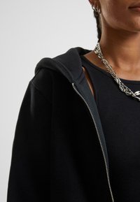 Felpa nera con zip realizzata in tessuto morbido, con una canotta a coste sotto. Caratterizzata da un cappuccio e dettagli metallici sulla zip.