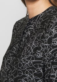 Blouse noire présentant un motif de lignes blanches ondulées, un col rond et des boutons sur le devant, confectionnée dans un tissu lisse.