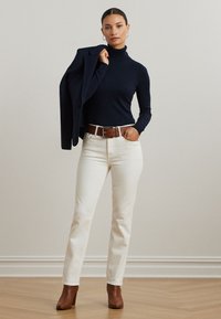Marinefarbener Rollkragenpullover, cremefarbene gerade Jeans, brauer Ledergürtel und braune Stiefeletten; die Person hält einen dunklen Blazer über der Schulter.