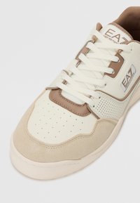 Tenisice s kremastim sintetičkim gornjim dijelom i taupe suede akcentima, perforiranim detaljima i bež gumiranim potplatom. Uključuju bijele vezice i brendiranje.