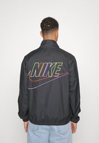 Svart vindjacka med hög krage, med en flerväg färgad "NIKE"-logotyp och swoosh-design på baksidan. Elastiska muddar och fåll.