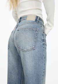 Jeans de mezclilla azul claro con cintura alta, con dos bolsillos traseros y un parche de cuero en la cinturilla. Textura suave, diseño de pierna recta.