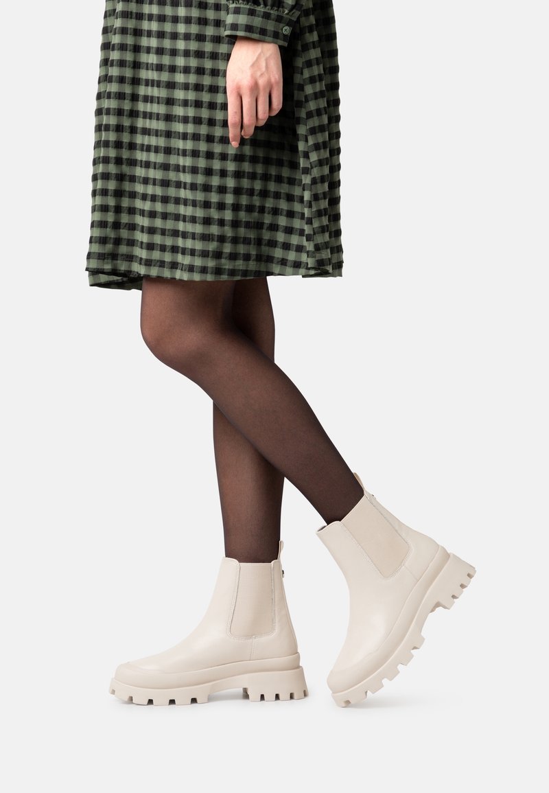 Bottes beige épaisses à la cheville avec des panneaux latéraux élastiques et une semelle texturée. Portées avec une robe à carreaux verte et des collants noirs.