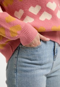 Pulover roz cu modele de inimi în alb și portocaliu. Manșetele și tivul cu nervuri, asortat cu blugi din denim de un albastru deschis. Prezentare a unei ținute casual.