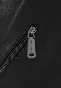 Black textured leather with a silver zip featuring a branded pull tab inscribed with "VALENTINO." Close-up view of the zipper detail.